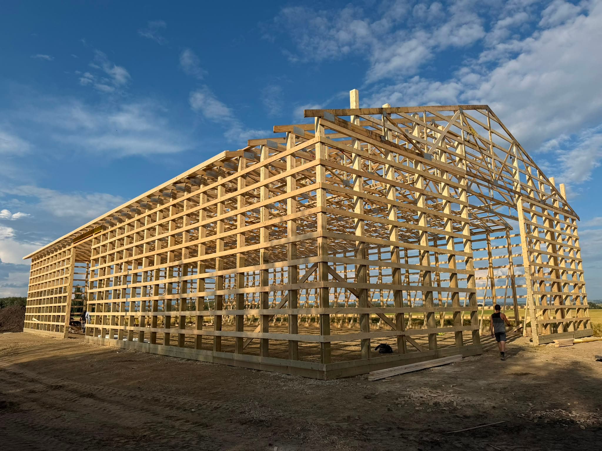 Wood post frame building under construction with exposed truss framing and a worker on site