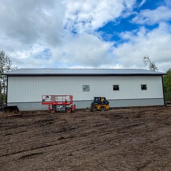 Completed post frame building with white metal siding and small windows near equipment on site
